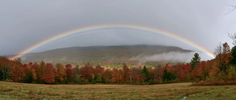 Double rainbow arching over autumn forest with misty mountains and colorful foliage in a rural meadow