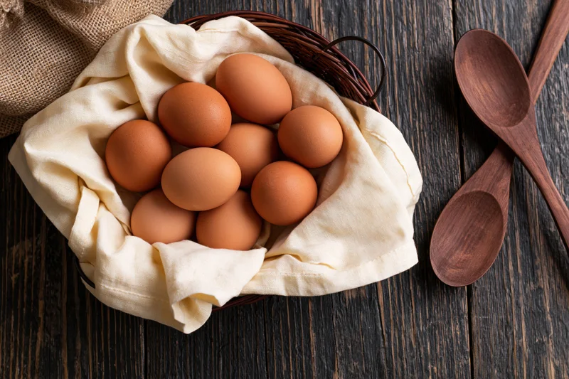 Brown eggs nestled in cream linen cloth inside a wicker basket on dark wooden table with wooden spoons beside