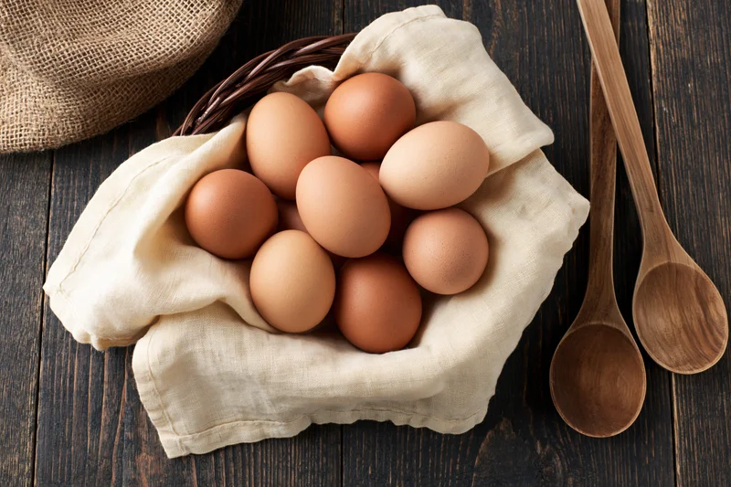 Brown eggs nestled in cream linen cloth inside a wicker basket on dark wooden table with wooden spoons beside