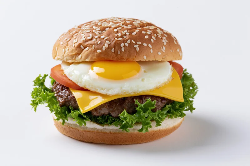 Beef cheeseburger topped with sunny-side-up fried egg and lettuce on sesame seed bun, isolated on white background