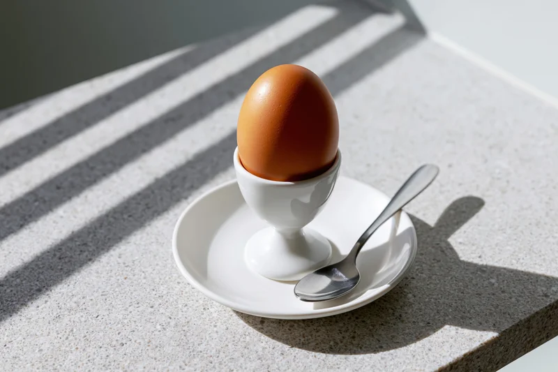 Brown boiled egg in white ceramic eggcup on plate with spoon on countertop in sunlight