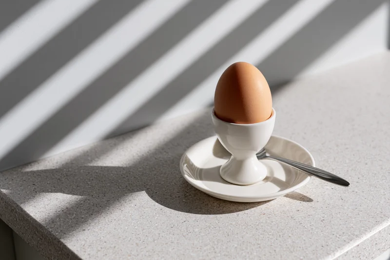 Brown boiled egg in white ceramic eggcup on saucer with spoon on countertop under striped sunlight
