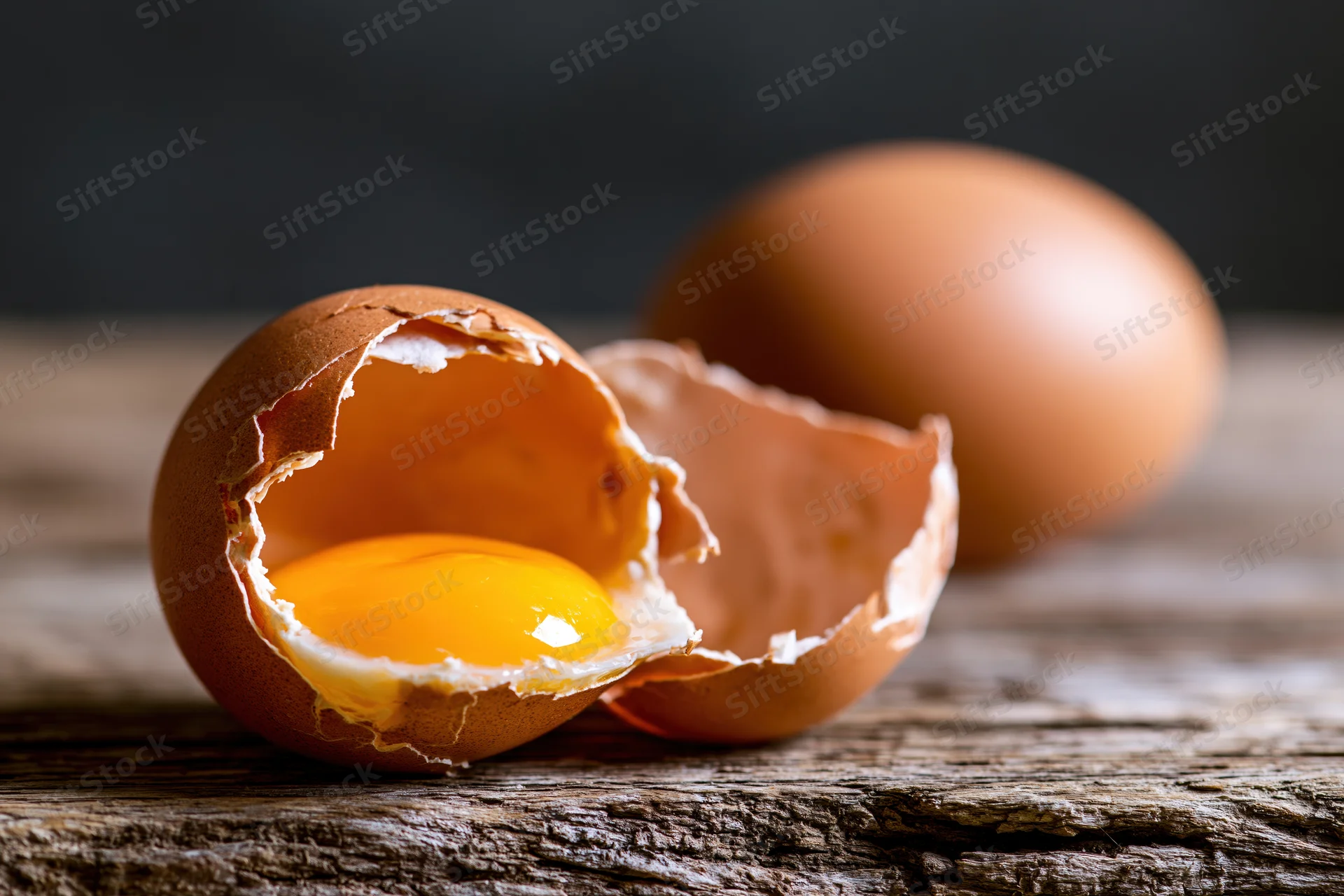 Cracked brown chicken egg split open revealing bright yellow yolk on wooden surface with whole egg blurred in background