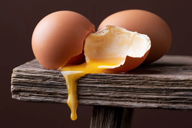 Cracked brown egg with yolk dripping off wooden table edge