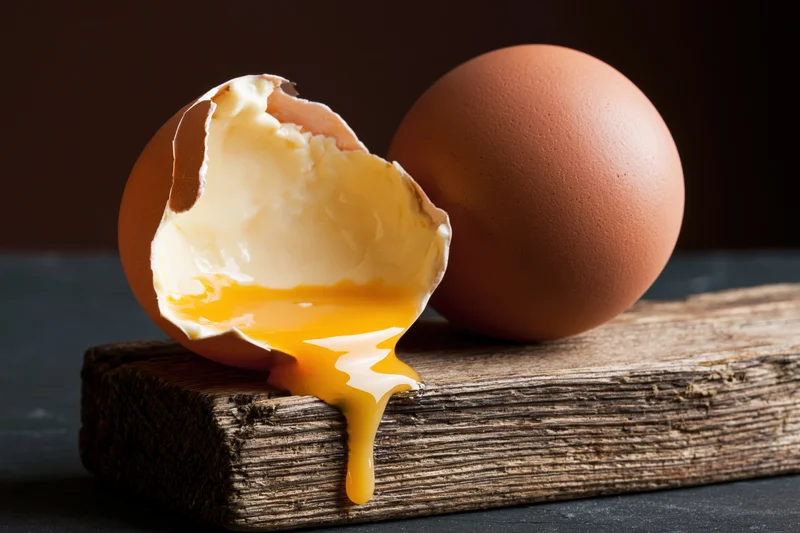 Cracked brown egg with yolk dripping off wooden board