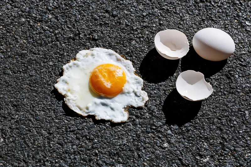 Fried egg cooking on hot dark asphalt pavement with cracked shells nearby