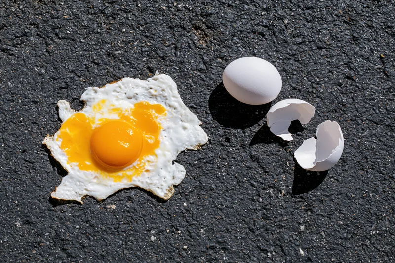 Fried egg on hot asphalt pavement with cracked shell nearby
