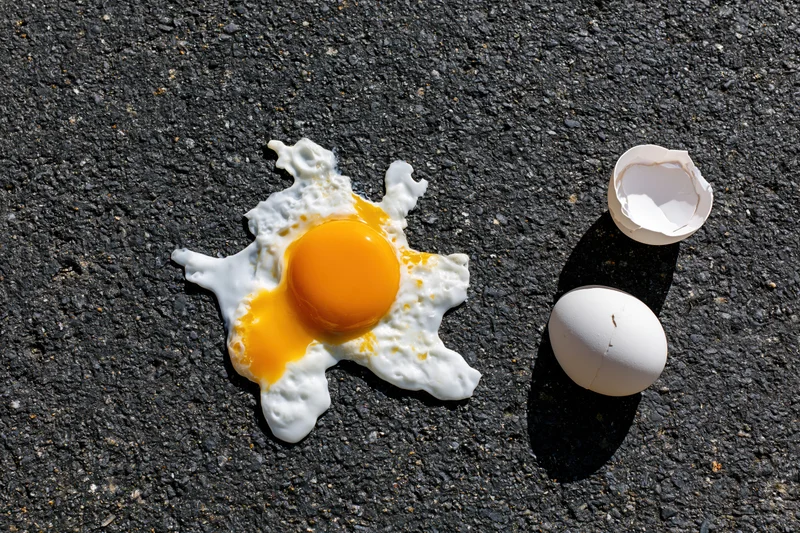 Fried egg on hot asphalt pavement with cracked shell and yolk visible under bright sunlight