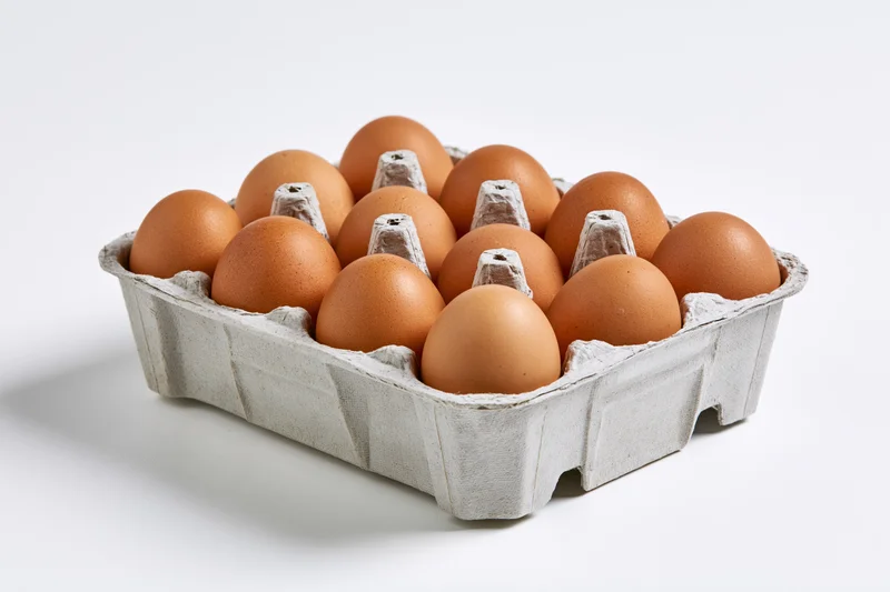 A dozen brown chicken eggs arranged in an open grey cardboard carton on a white surface