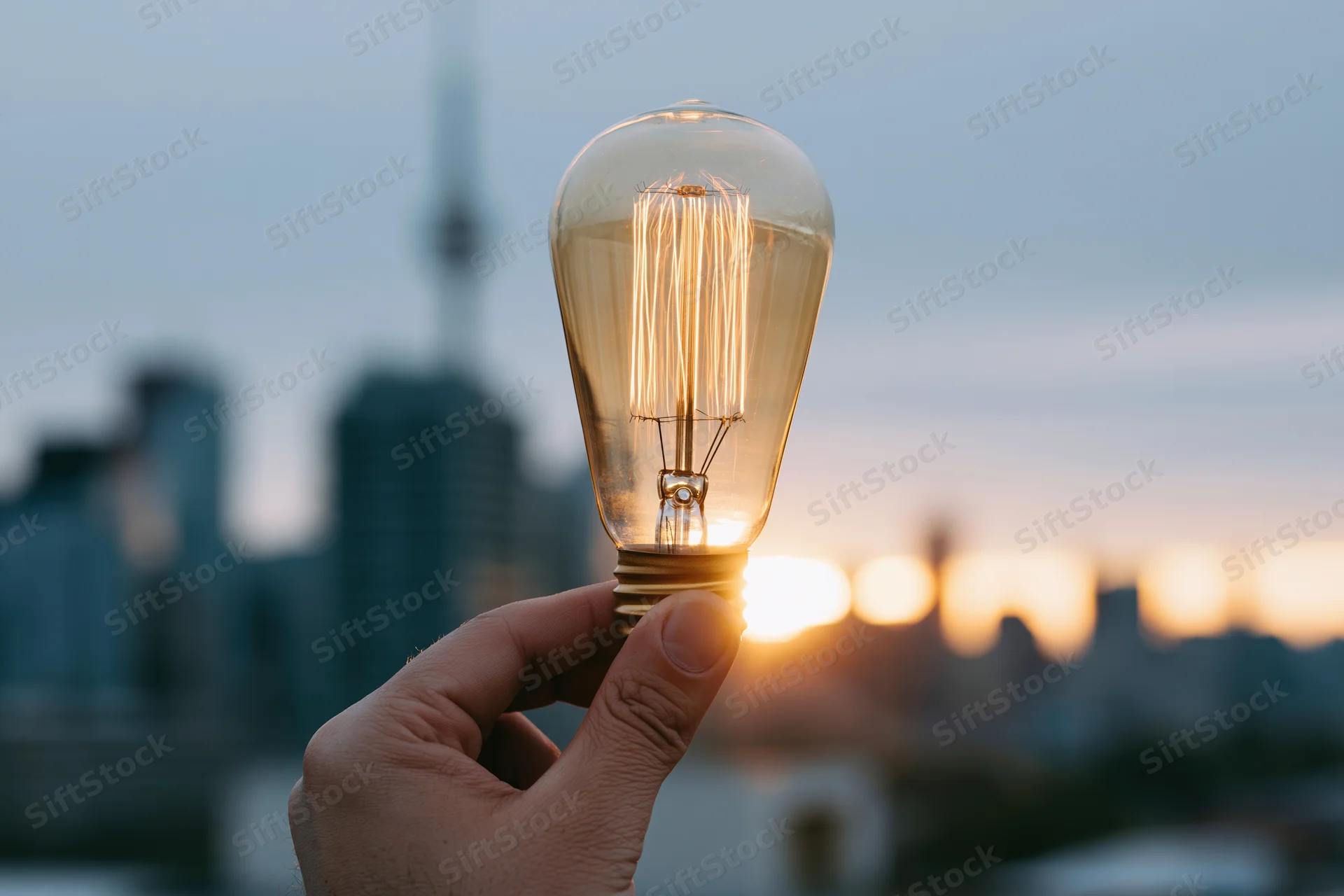 Hand holding glowing edison filament lightbulb against city skyline at sunset