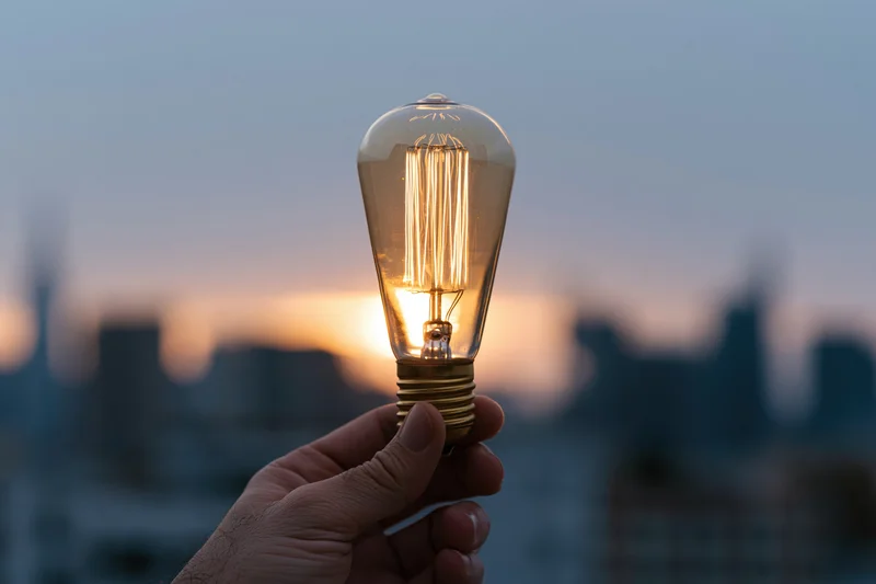 Hand holding glowing edison filament lightbulb against blurred cityscape at sunset
