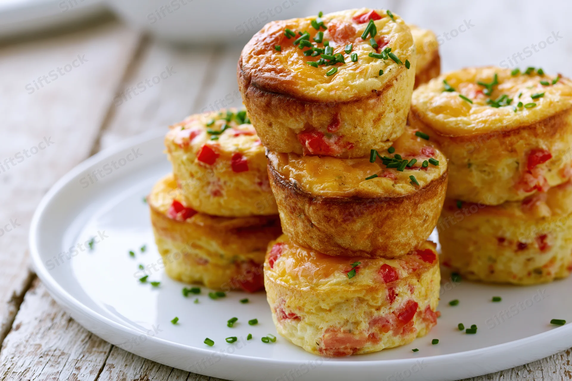 Golden-brown egg muffin cups stacked on a white plate with chives and red bell pepper bits, set on a rustic wooden table