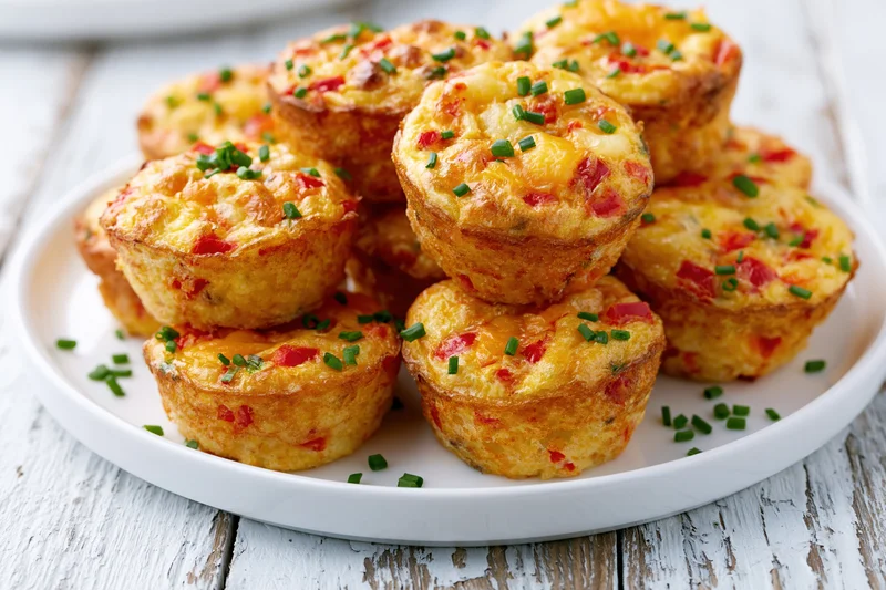 Golden-brown egg muffin cups filled with red bell pepper and cheese, garnished with chives, piled on a white plate on a rustic wooden table
