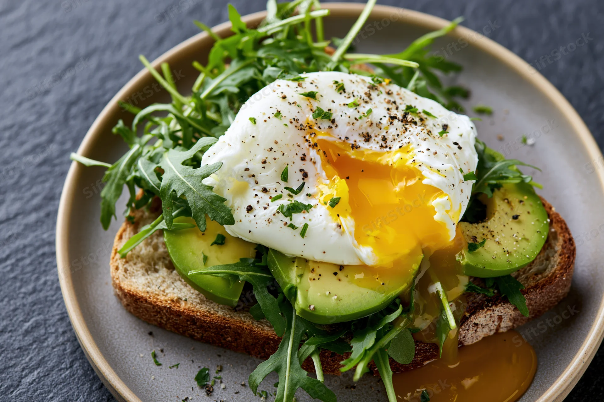 Poached egg with broken yolk running over avocado toast on a plate with arugula and herbs