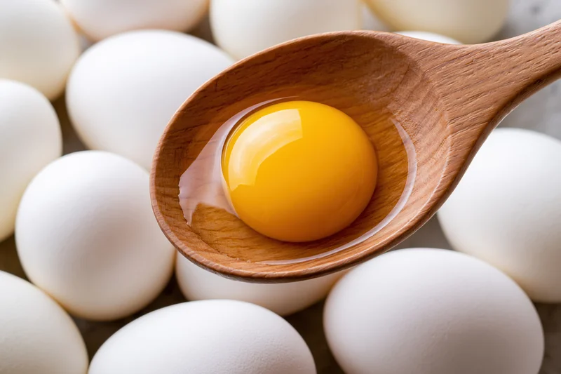 Raw egg yolk with clear albumen resting in a smooth wooden spoon surrounded by whole eggs