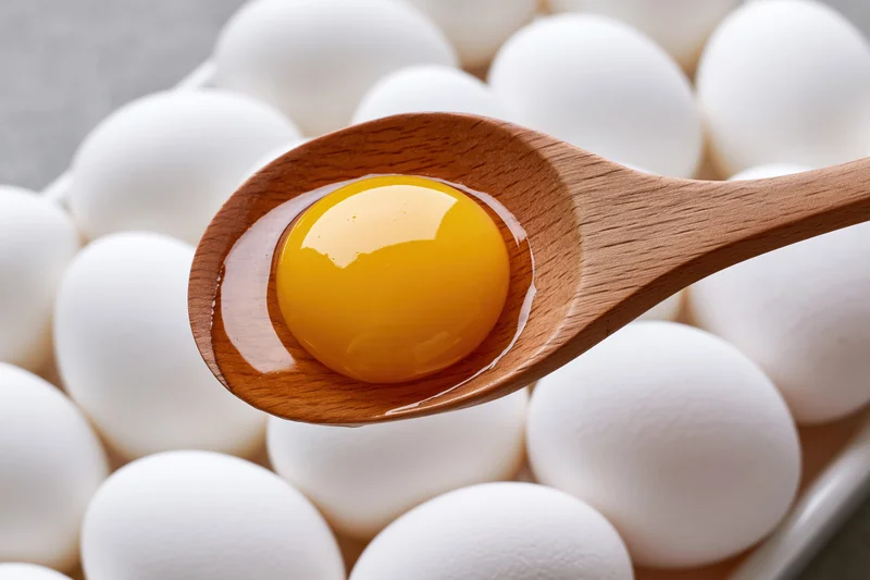 Raw egg yolk with clear albumen resting in a wooden spoon above a tray of white eggs