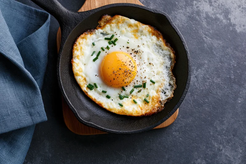 Sunny-side-up fried egg in seasoned cast iron skillet on wooden trivet with blue cloth napkin beside