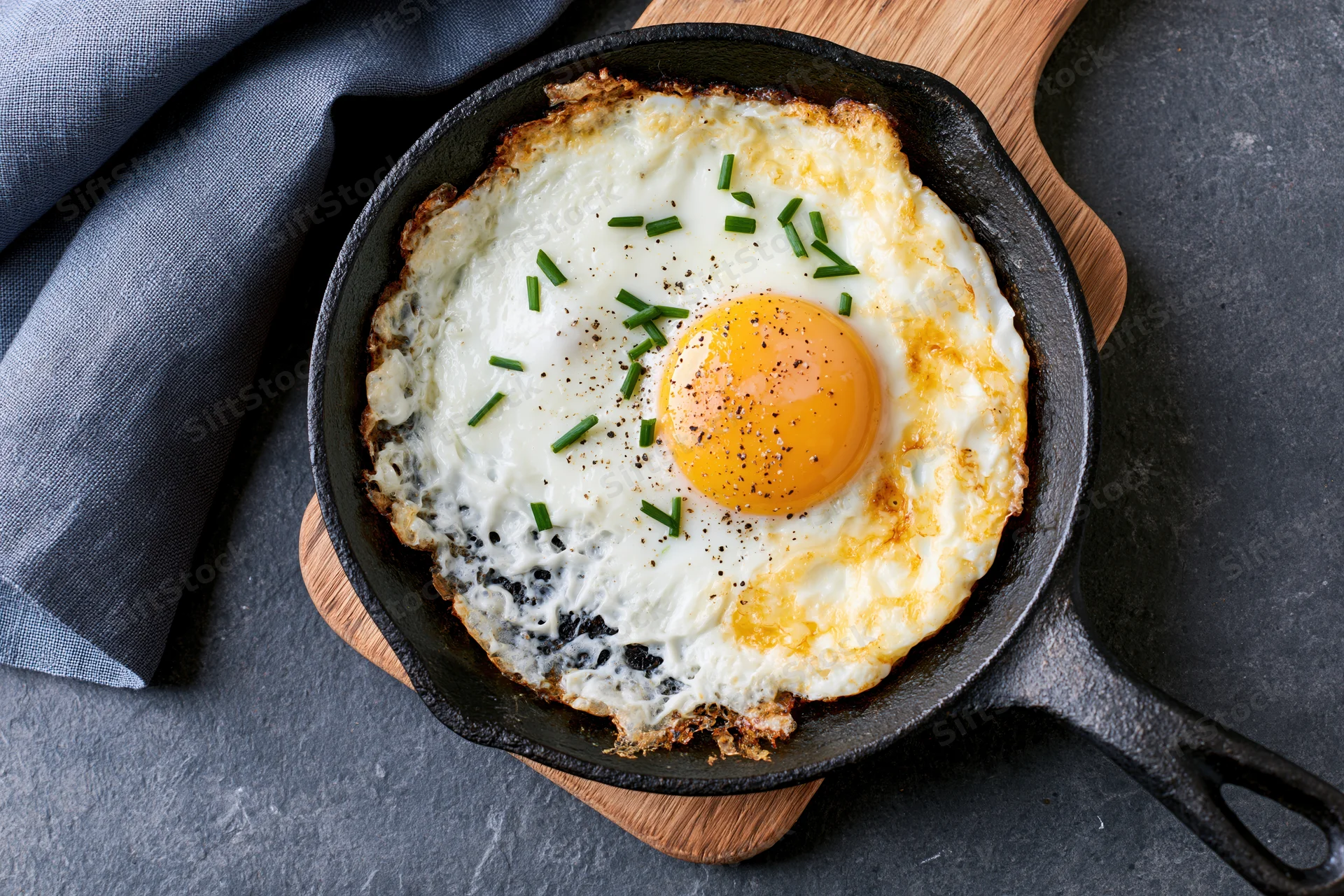 Sunny-side-up fried egg in a seasoned cast iron skillet on a wooden trivet with chives and pepper sprinkled on top