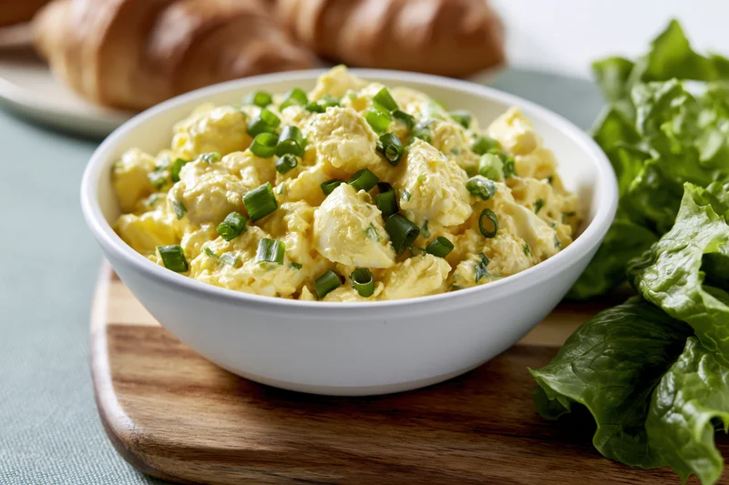 Chunky egg salad in a white ceramic bowl garnished with green onions on a wooden board with lettuce and croissants nearby