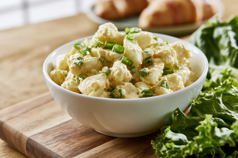 Chunky egg salad in a white ceramic bowl on a wooden cutting board with lettuce and croissants nearby