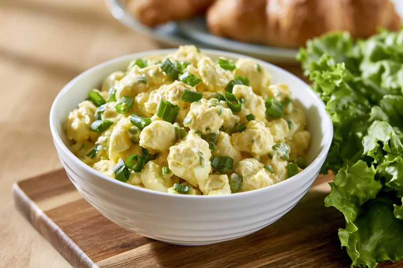 Chunky egg salad in a white ceramic bowl on a wooden cutting board with fresh lettuce and croissants in the background