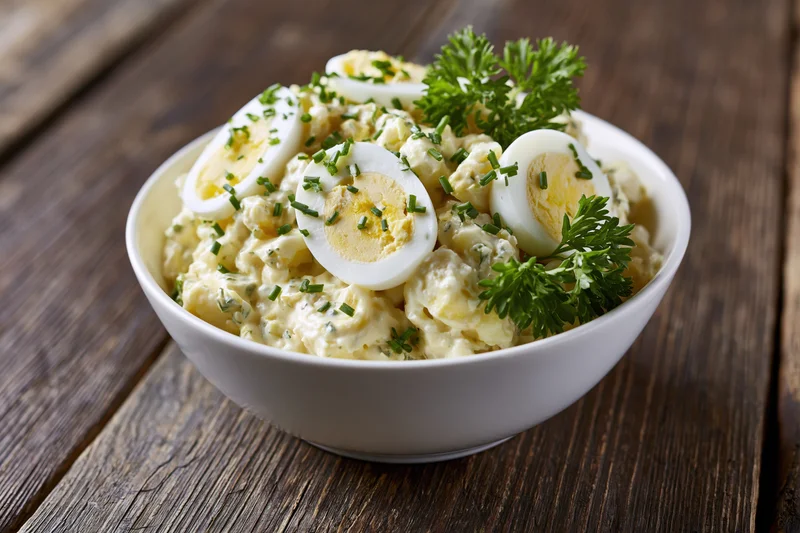 Creamy egg salad in a white ceramic bowl topped with sliced hardboiled eggs and fresh herbs on a wooden table