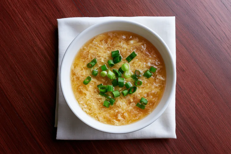 Egg drop soup in a white ceramic bowl garnished with green onions on a wooden table with a cloth napkin