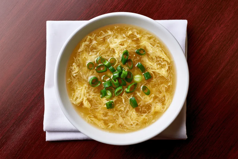 Egg drop soup in a white ceramic bowl garnished with green onions on a dark wooden table with a folded white napkin beneath