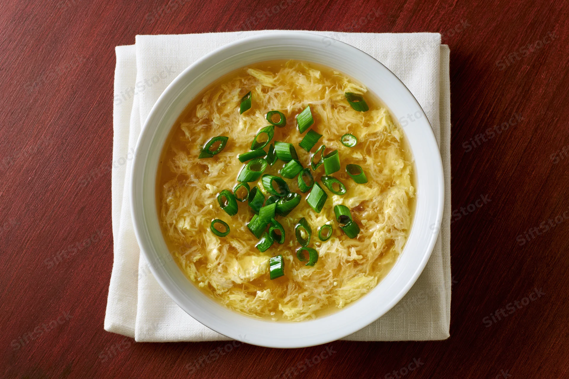 Egg drop soup in a white ceramic bowl garnished with green scallions on a wooden table with a beige napkin