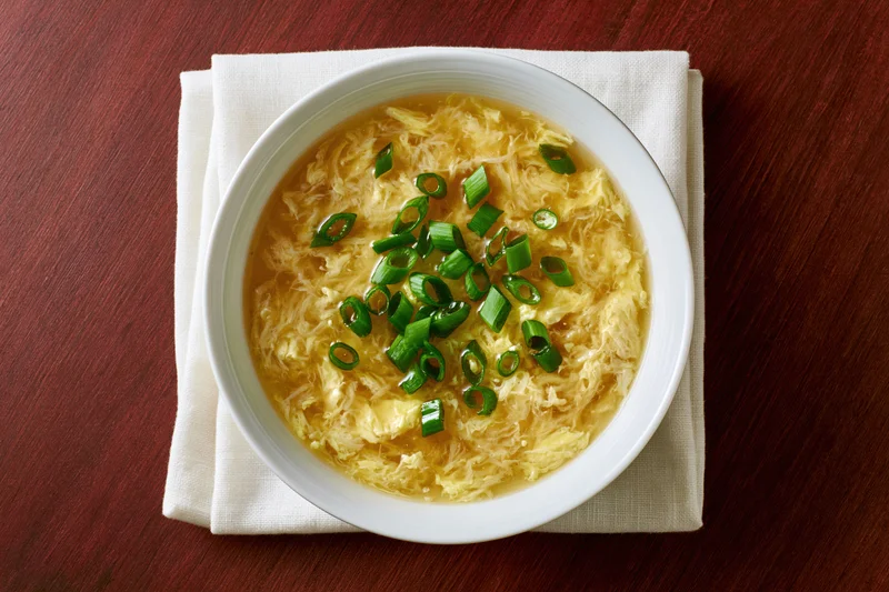 Egg drop soup in a white ceramic bowl garnished with green scallions on a wooden table with a beige napkin