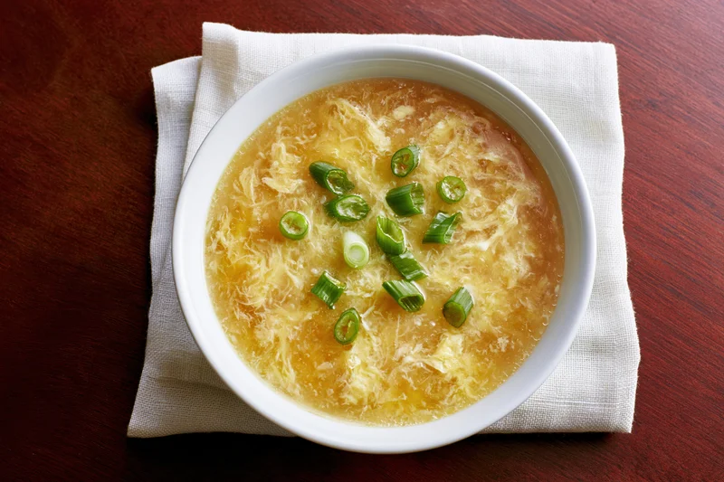 White ceramic bowl of egg drop soup with wispy cooked egg and green onion garnish on wooden table with linen napkin