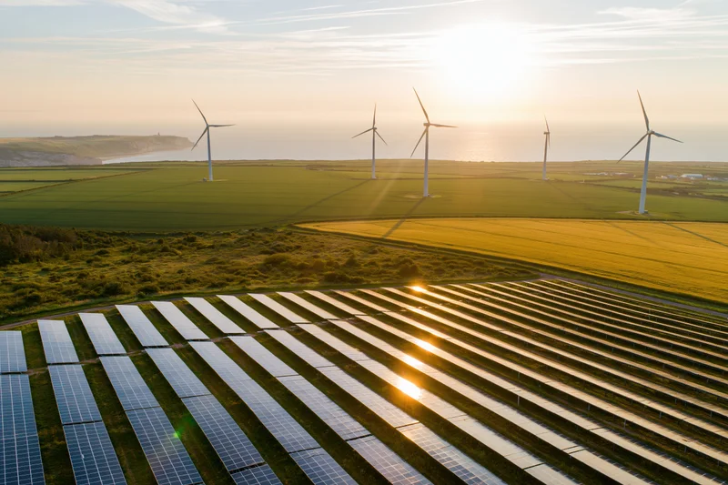 Solar panels in rows across a green field with wind turbines in the distance near the coast at sunset