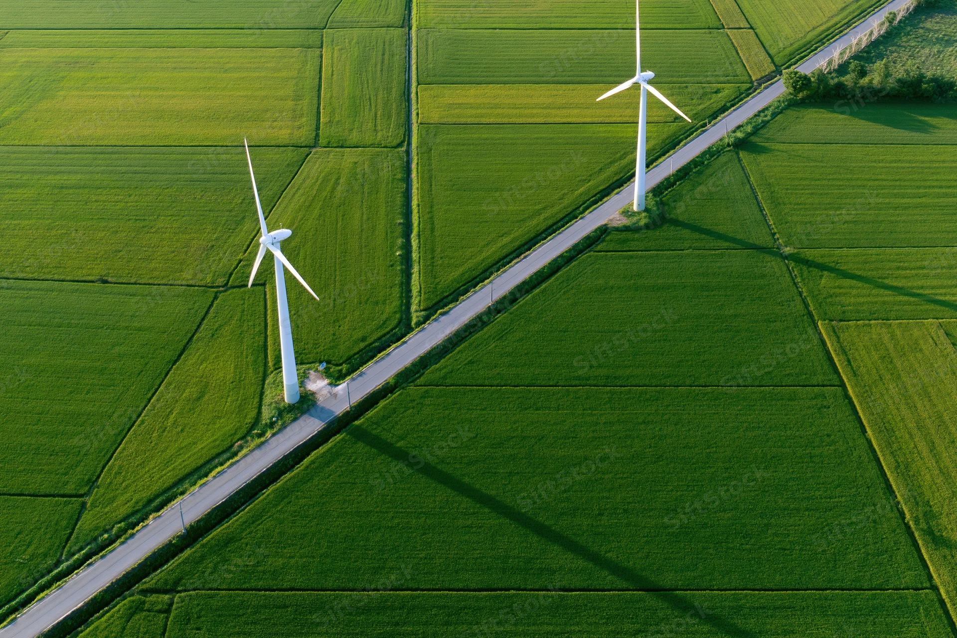Two white wind turbines standing in a green agricultural field viewed from above along a narrow road