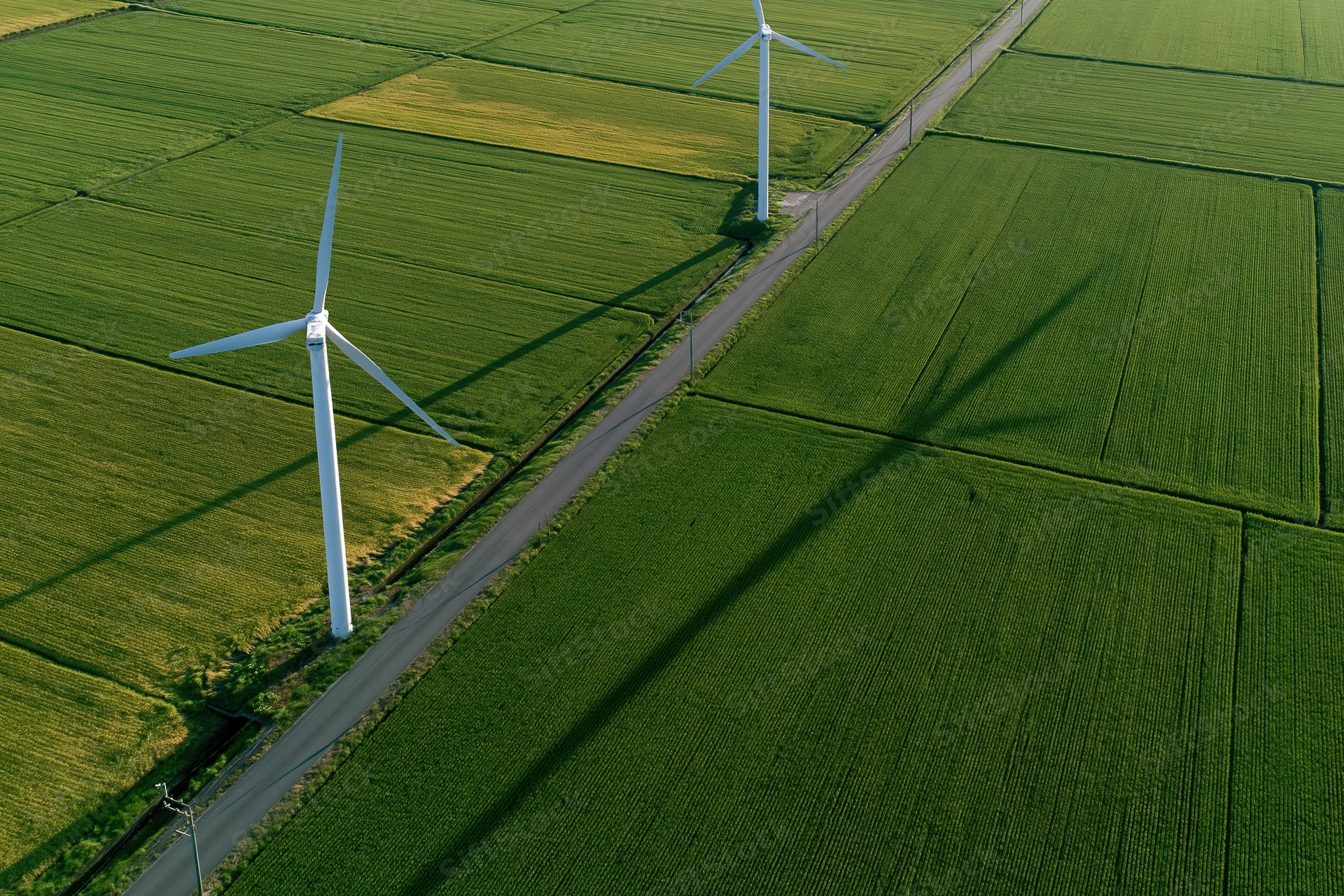 Two white wind turbines standing in a green agricultural field viewed from above along a narrow road