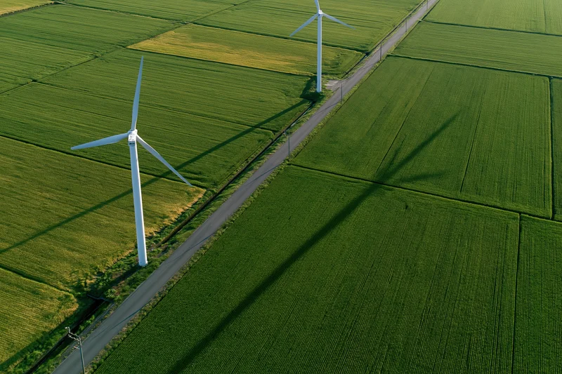 Two white wind turbines standing in a green agricultural field viewed from above along a narrow road