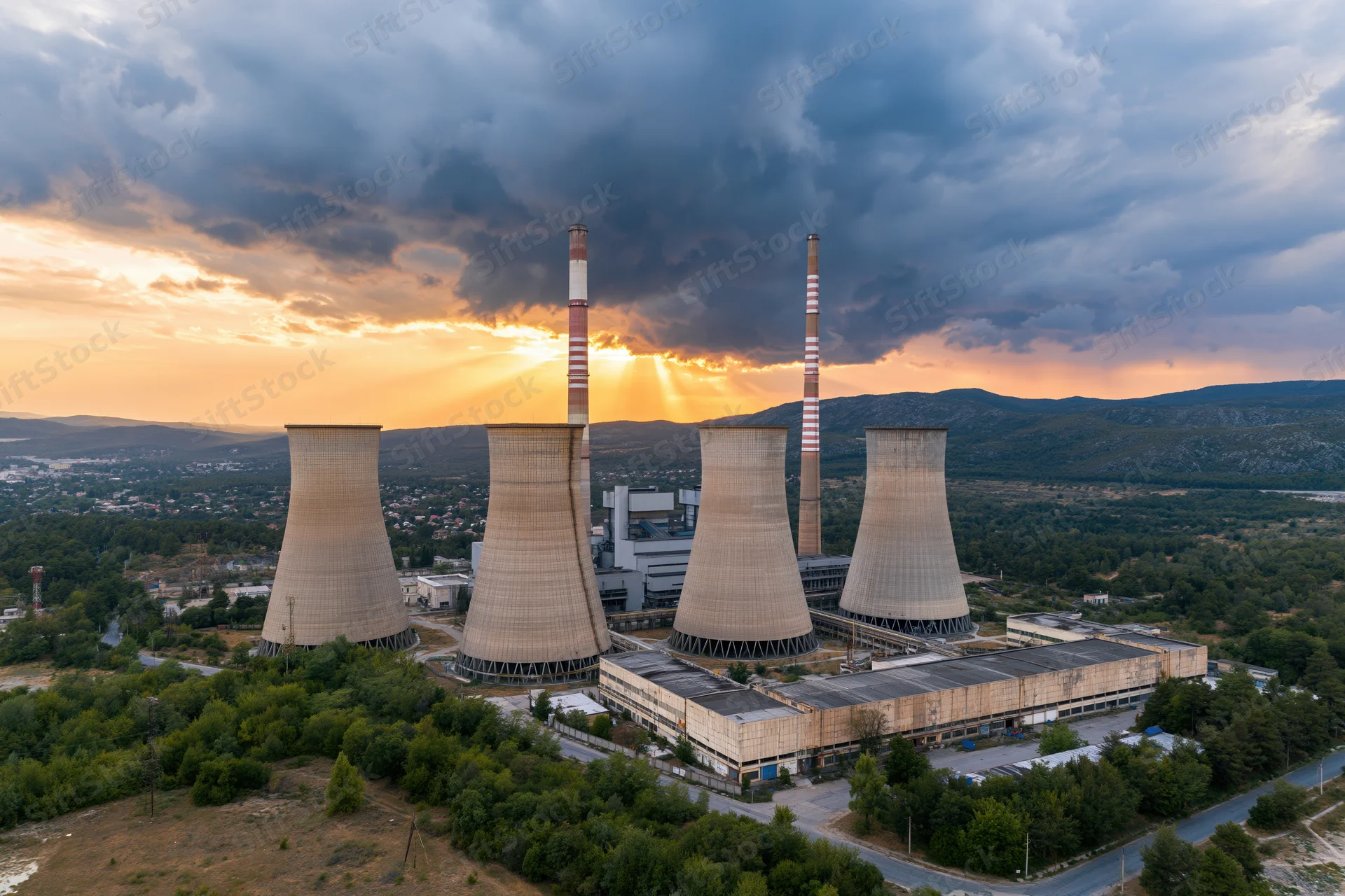 Aerial view of a coal-fired power plant with cooling towers and smokestacks at sunset surrounded by trees and hills