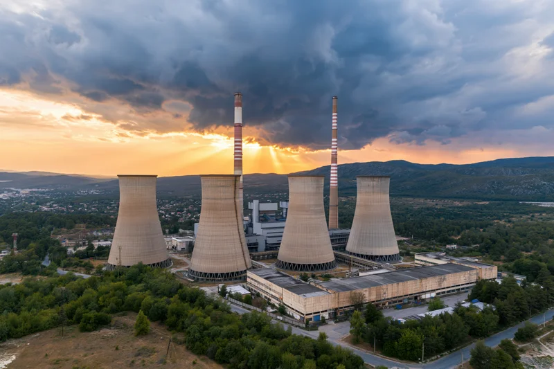 Aerial view of a coal-fired power plant with cooling towers and smokestacks at sunset surrounded by trees and hills