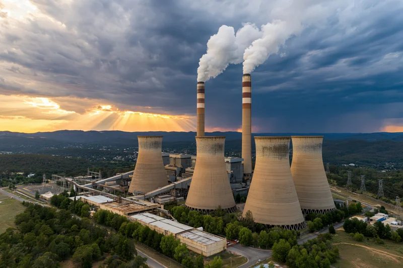 Aerial view of a coal thermal power plant with cooling towers and smokestacks emitting steam against a cloudy sunset sky