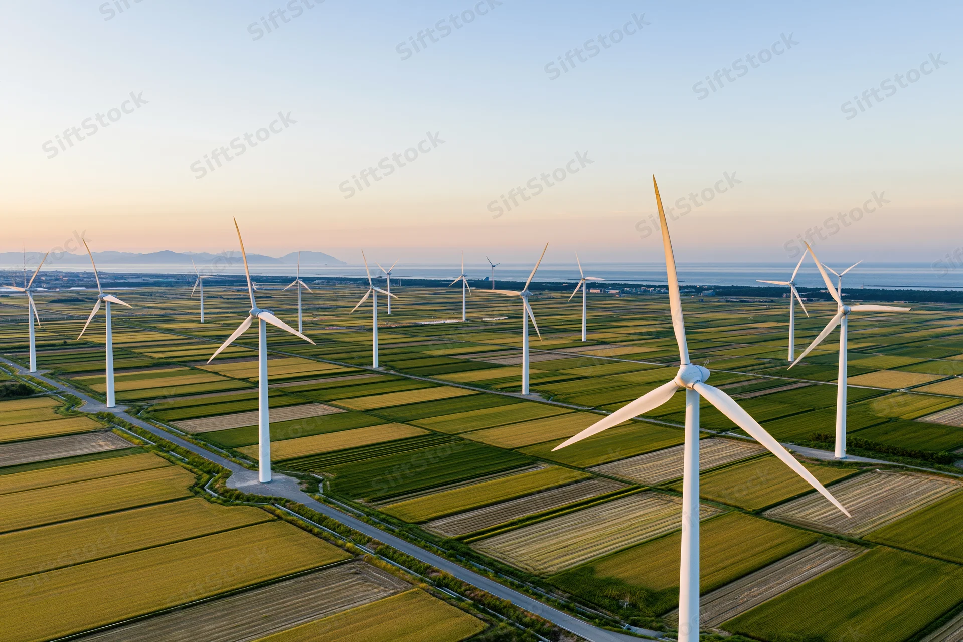 Wind turbines standing across flat green farmland near coast at sunset