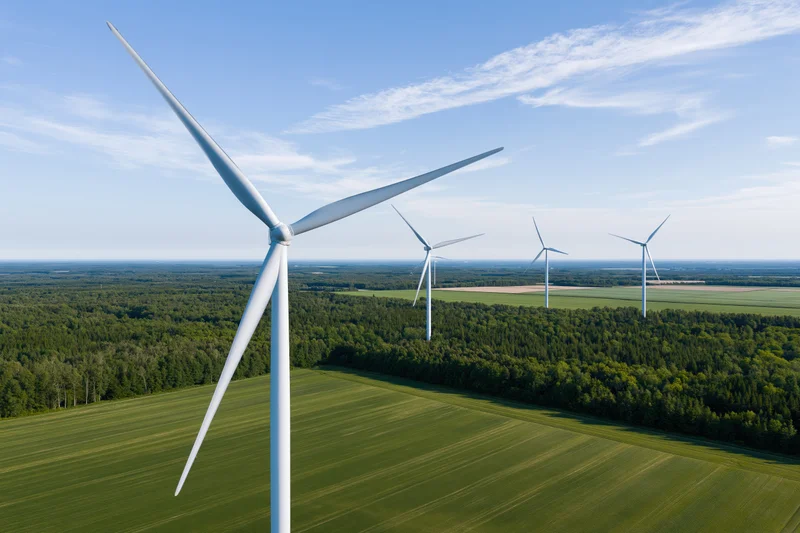 Wind turbines in a row across green fields and forest under blue sky from aerial view