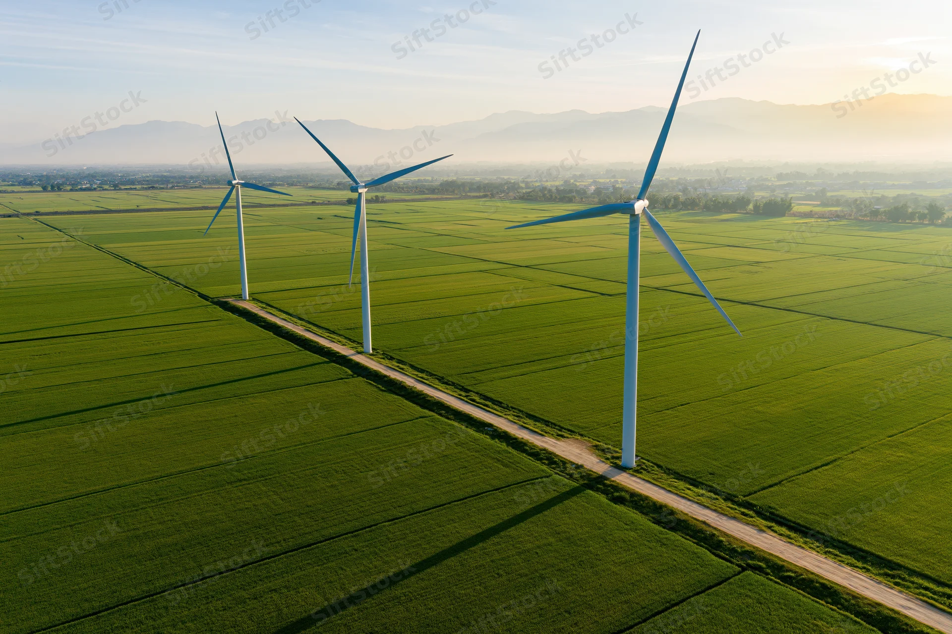 Three wind turbines aligned along a dirt road in a green agricultural field with mountains in the distance at sunset