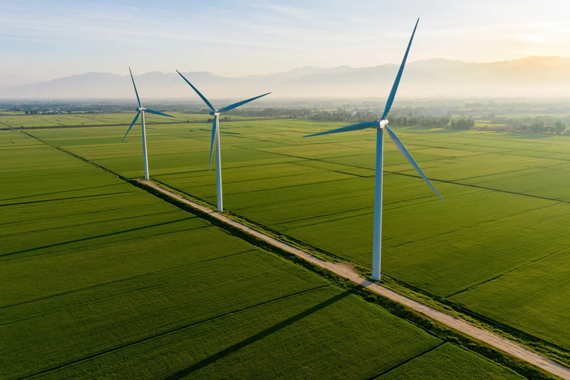 Three wind turbines aligned along a dirt road in a green agricultural field with mountains in the distance at sunset
