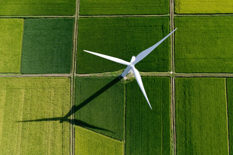 Single white wind turbine casting long shadow over green crop fields from top-down aerial view