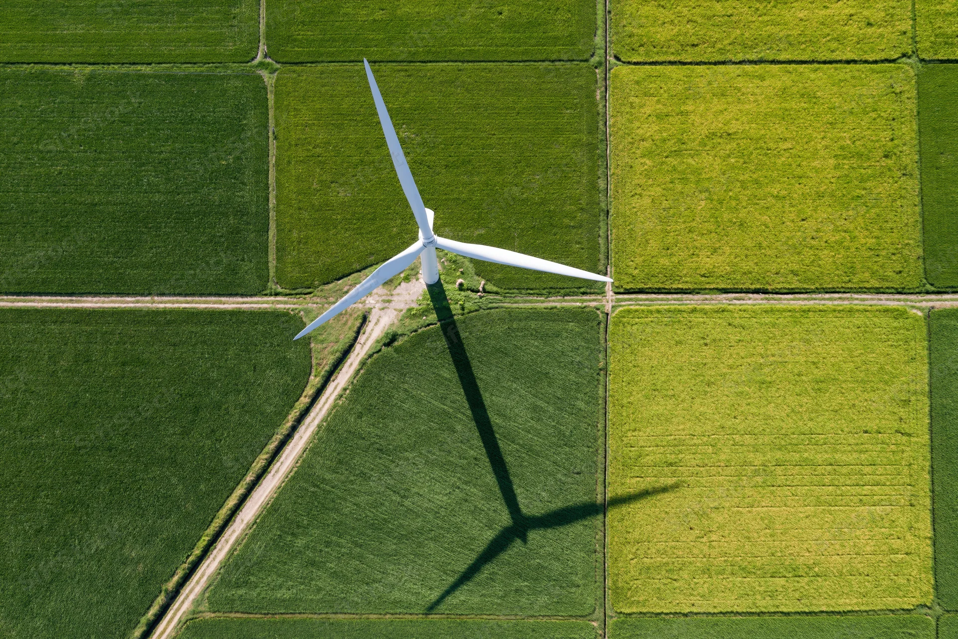 Single white wind turbine casting a long shadow over a grid of green and yellow crop fields from above
