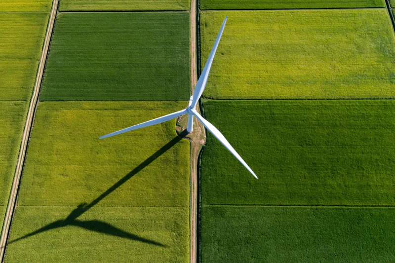 Single white wind turbine casting long shadow on green agricultural fields from top-down aerial view