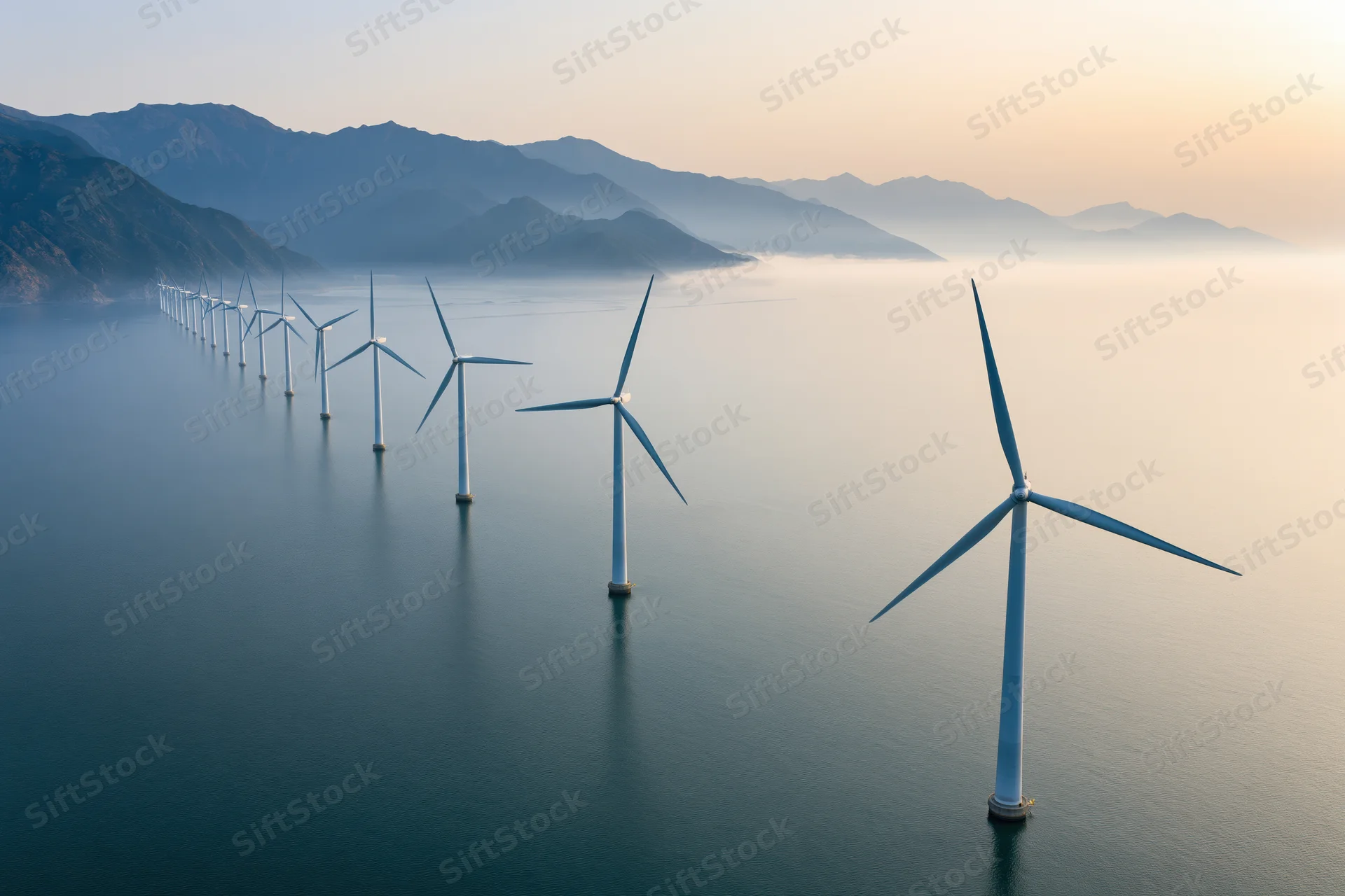 Row of offshore wind turbines standing in calm ocean water near mountains at sunrise