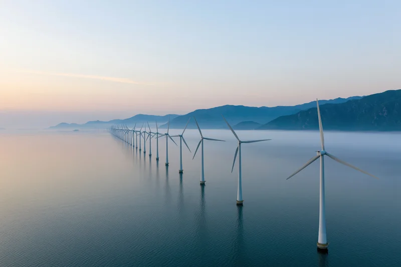 Row of offshore wind turbines aligned in calm ocean waters near coastal mountains at twilight