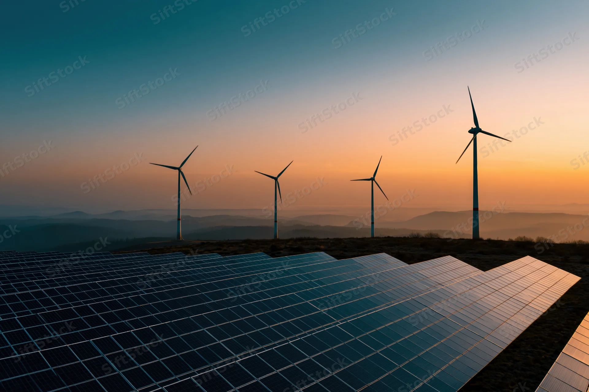 Solar panels arranged in rows with wind turbines on a hill at sunset
