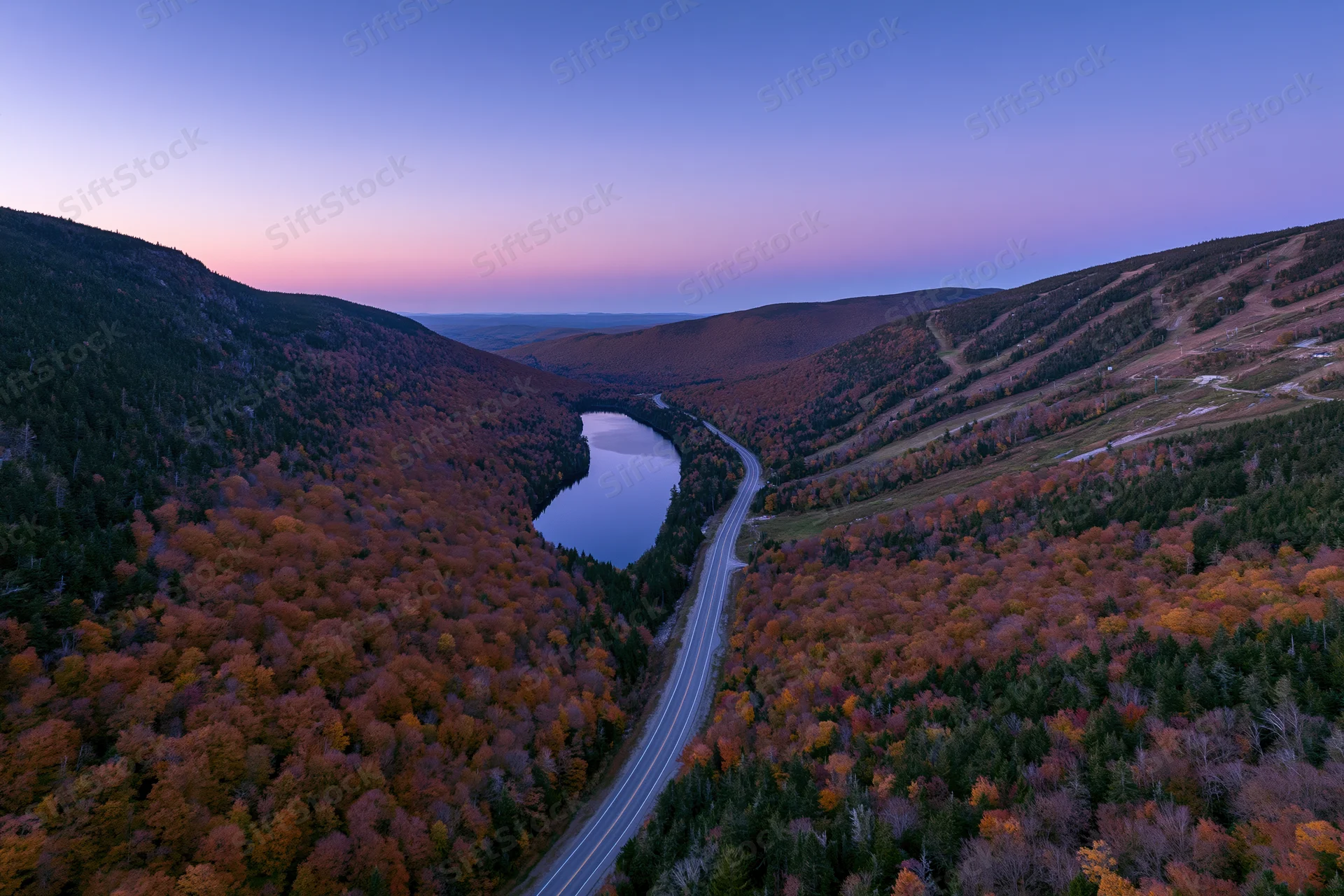 Aerial view of a winding road through a mountain valley with autumn foliage and a lake at dusk