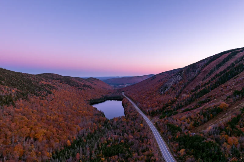 Aerial view of a winding road through a mountain valley with autumn foliage and a lake at dusk